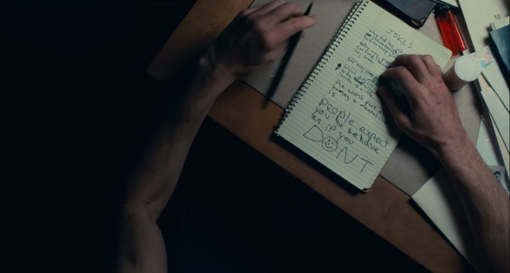 Mãos escrevendo em um caderno sobre a mesa. Há remédio, papeis, isqueiro na mesa. 