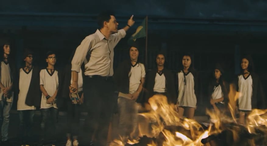 Professor queimando livros e rodeado por crianças com capuzes em Escola Sem Sentido. 