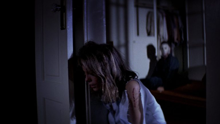 Mulher encostada na batente de uma porta enquanto um homem se levanta atrás dela