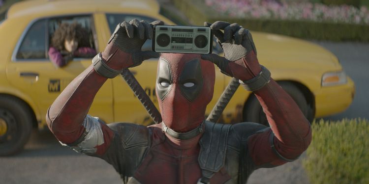 Deadpool esta segurando uma caixa de som olhando para frente, com um taxi ao fundo. 