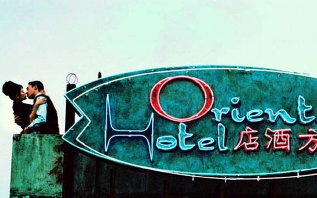 Imagem do filme 2046, onde um casal, composto por um homem e uma mulher, se beijam em cima do telhado de um hotel, cujo letreiro em neon, nas cores azul e vermelho, dizem "Oriental Hotel".