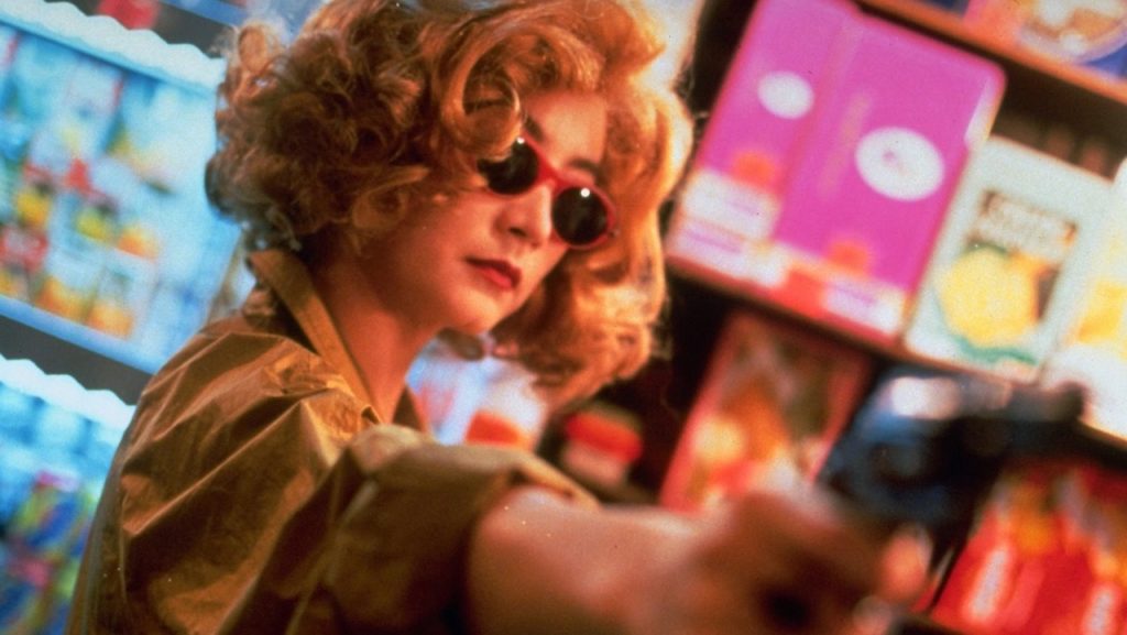 Imagem do filme Chungking Express, em que uma mulher, retratada em ângulo holandês inclinado para a direita, com óculos de sol de armação vermelha em destaque, aponta uma arma para algo ou alguém situado do lado direito da câmera. Diretor Wong Kar Wai