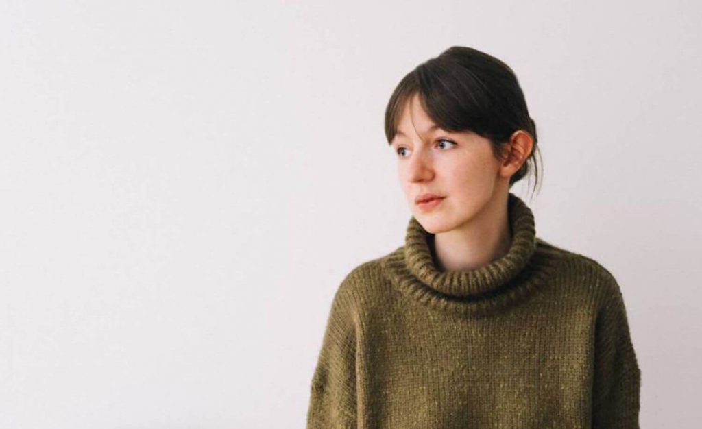 A escritora Sally Rooney, autora de Pessoas Normais, vestida com um suéter verde musgo que contrasta com a parede branca. Otageek