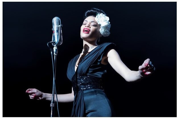 Vestida de preto com uma flor no cabelo, a cantora Andra Day interpretando Billie Holiday. Otageek