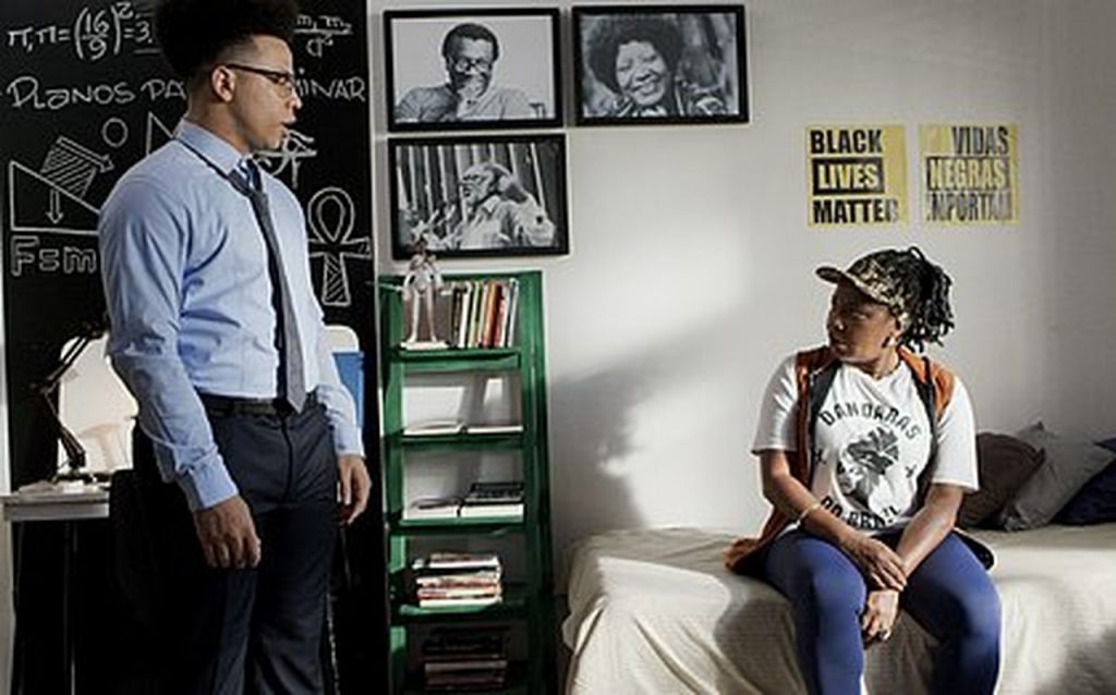 Um homem em pé ao lado de uma instante cheia de objetos, e uma mulher sentada na cama