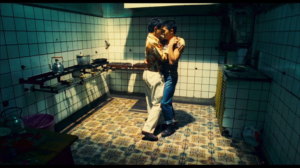 Imagem do filme Happy Together, onde um casal, constituído por dois rapazes, dança abraçado exatamente no centro de uma cozinha em condições precárias. Toda a cozinha está na penumbra, exceto pela parte central do plano, onde o teto está aberto e a luz do sol ilumina o casal. Diretor Wong Kar Wai.