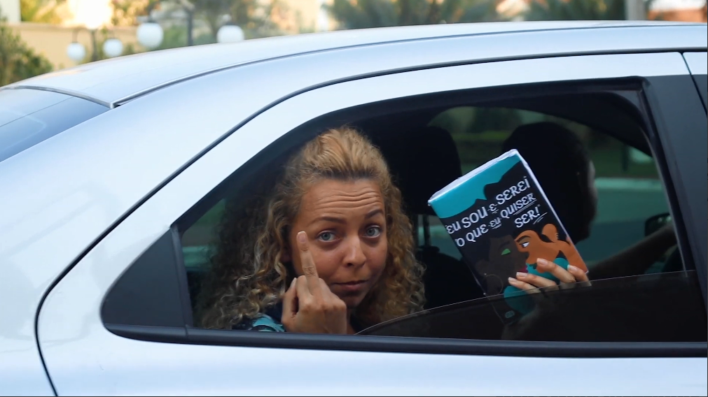 A protagonista dá dedo de dentro de um carro enquanto mostra seu livro, Eu sou e serei o que eu quiser ser