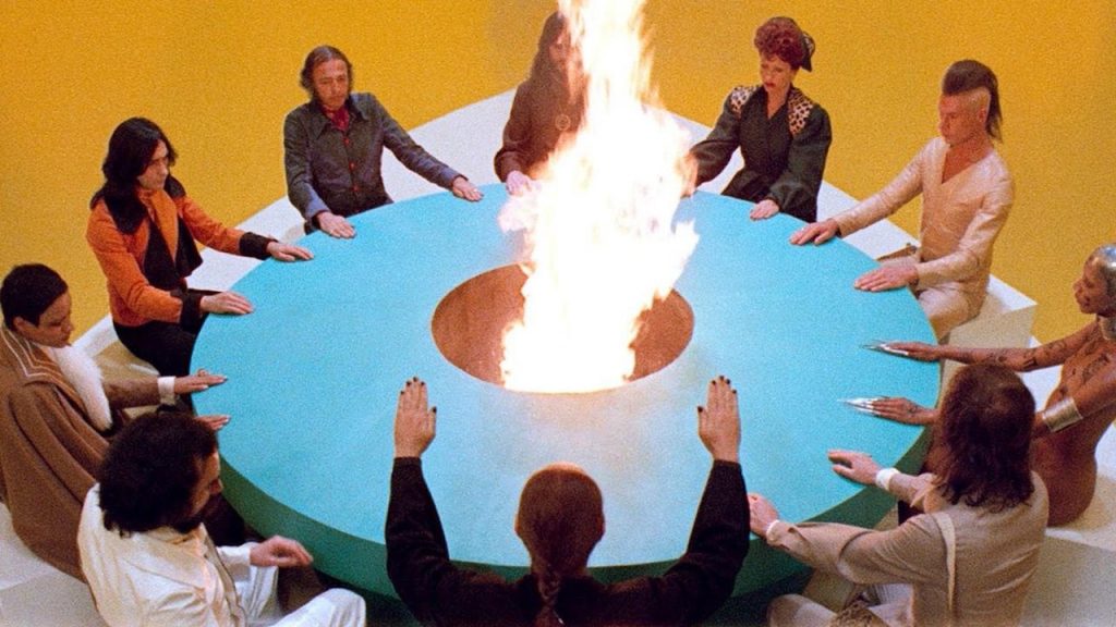 Imagem do filme A Montanha Sagrada (1973). Nela, dez pessoas estão sentadas à uma távola redonda azul, com um fogaréu no centro da távola. 