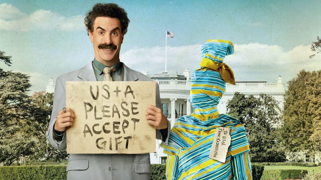 Imagem promocional do filme Fita de Cinema Seguinte de Borat. Nela, vemos o autointitulado repórter do Cazaquistão na frente da Casa Branca segurando uma placa, na qual se lê "US+A POR FAVOR ACEITEM PRESENTE". Do lado dele, vê-se um embrulho azul e amarelo em forma de pessoa, com uma etiqueta escrita "PARA: US+A / DE: CAZAQUISTÃO"