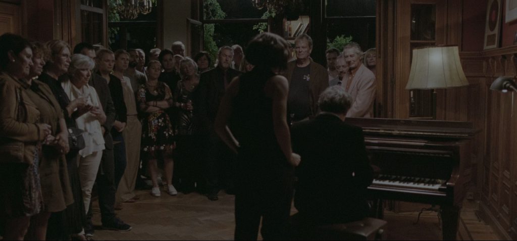 Cena do terceiro ato do filme, em que Lou (Olivia Cooke) e seu pai (Mathieu Amalric), ambos vestidos em roupas pretas, tocam uma música para convidados de sua festa. Ambos estão de costas para a câmera, enquanto o público está de frente. Lou está de pé, cantando, enquanto o pai está sentado, tocando em um piano. No sentro do público, em sua maioria pessoas de meia-idade, vemos Ruben, trajando uma camiseta cinza e parecendo deslocado naquele evento.