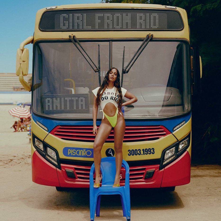 Anitta em pé em cima de uma cadeira de plástico azul, em fente a um ônibus que tem escrito em cima Girl From Rio, e Anitta no vidro frontal do ônibus. O fundo é uma praia do Rio de Janeiro.