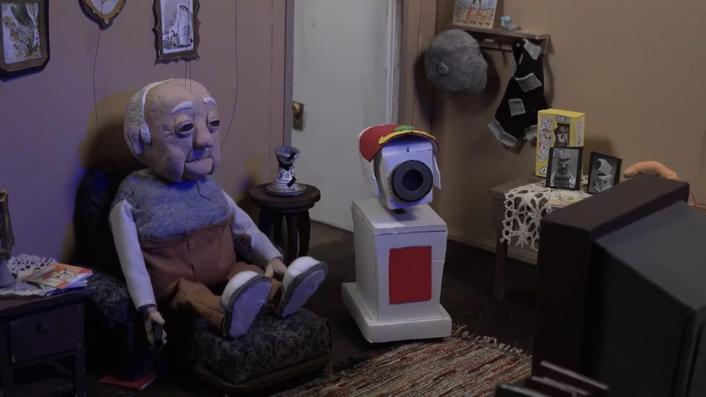 Mr. Nakashima assiste TV em companhia de se robô, trajando um chapéu. 