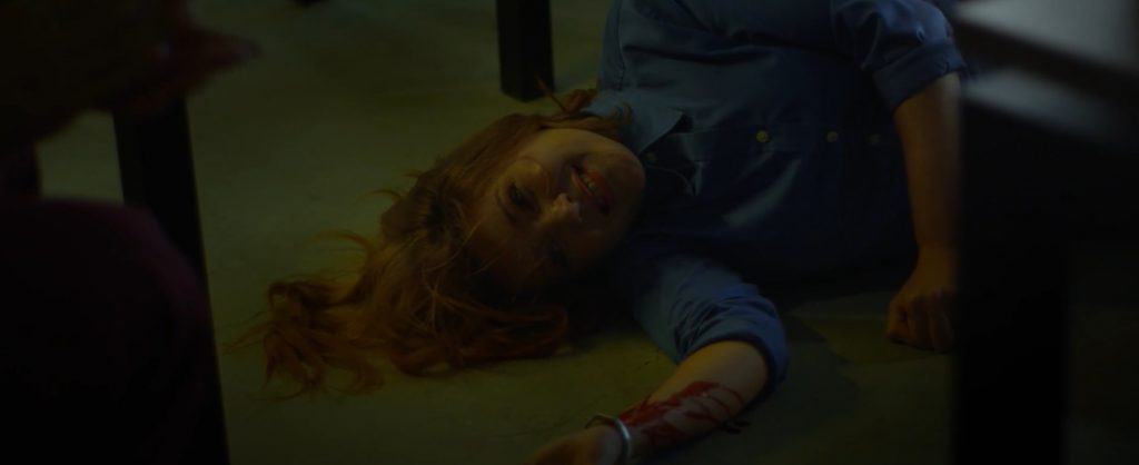 Personagem deitada no chão - morta - com um sorriso no rosto e o pulso direito sangrando.