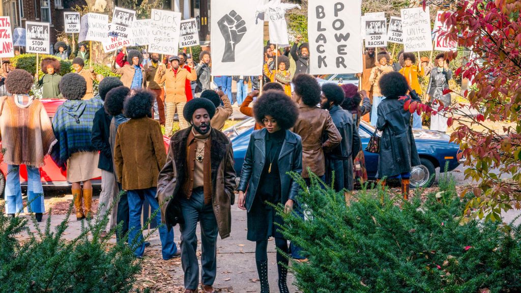 homem e mulher negros conversando, em roupas de couro, cabelos black power. atrás, grupo de pessoas negras protestando