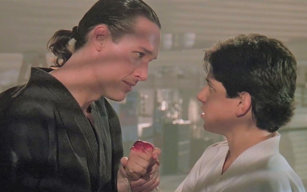 Personagem Terry Silver e Daniel Larusso em cena do filme Karate Kid 3. Terry está segurando o braço de Daniel. Daniel tem a mão machucada sangrando, e os dois estão se encarando.