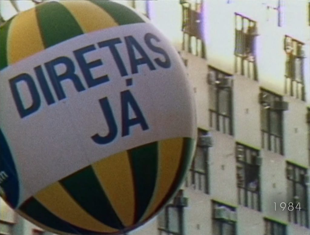 Balão com as listras verde-amarelo escrito DIRETAS JÁ em caixa alta. a data 1984 aparece no canto inferior direito da tela no documentário O Despertar da Florestania 