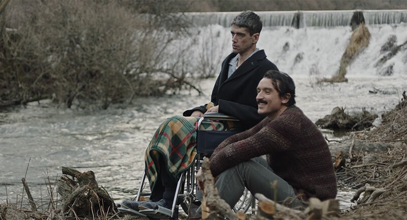 Imagem do filme do Cinefantasy: Amigo, de Oscar Martin. Na cena temos dois rapazes sentados perto de um rio em um dia nublado e frio. Um rapaz está na cadeira de rodas com um olhar bravo e o outro sentado no chão está sorrindo