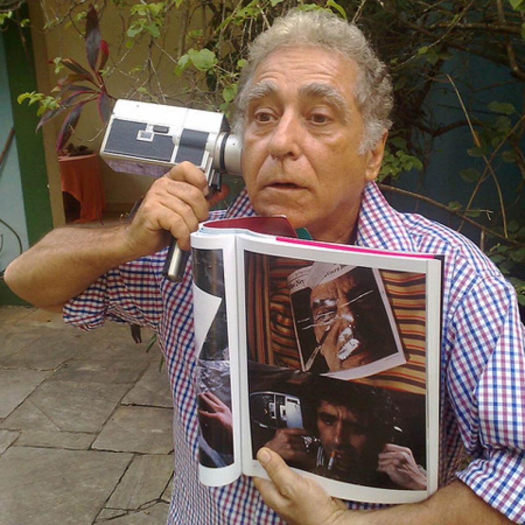 Foto do cineasta Neville D'almeida, que participou do Cinefantasy segurando com a mão direita uma camera encostada em seu rosto e com a mão esquerda um livro com figuras de cenas de filmes.