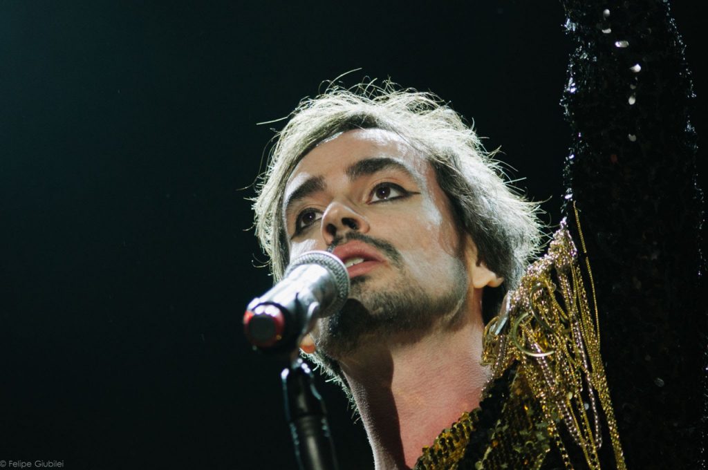 O cantor Johnny Hooker no palco, em close, na frente de um fundo preto, trajando preto também, com detalhes em dourado na roupa. Johnny tem cabelos que vão até o ombro, barba baixa, e usa maquiagem nos olhos.