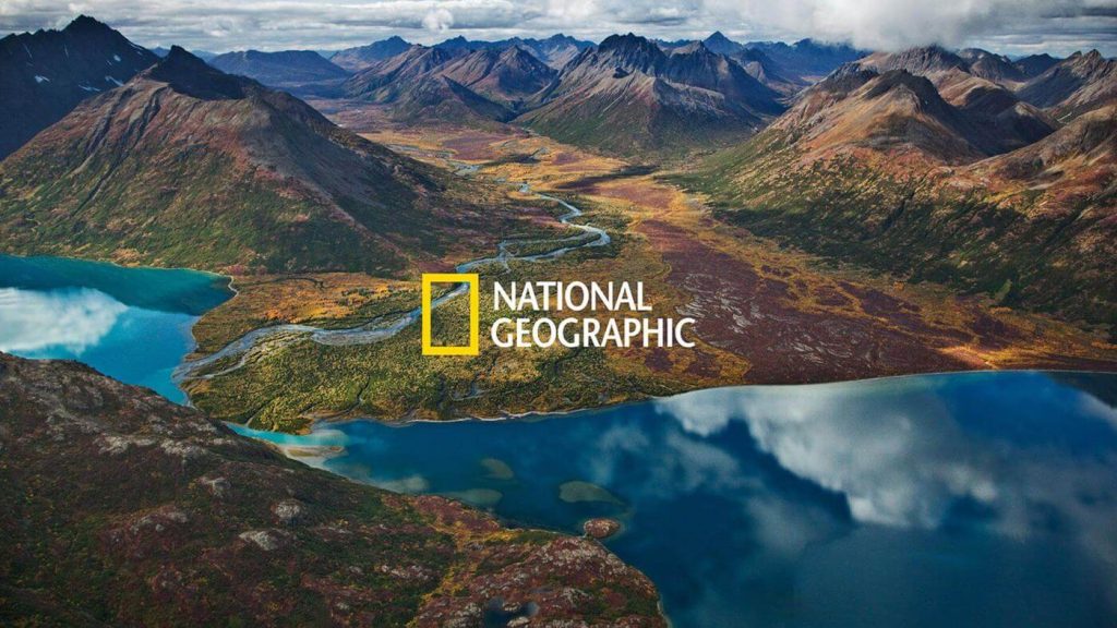 A imagem mostra uma cadeia de montanhas com um rio passando ao meio. As nuvens estão refletidas no rio e centralizado está o logo do National Geographic.