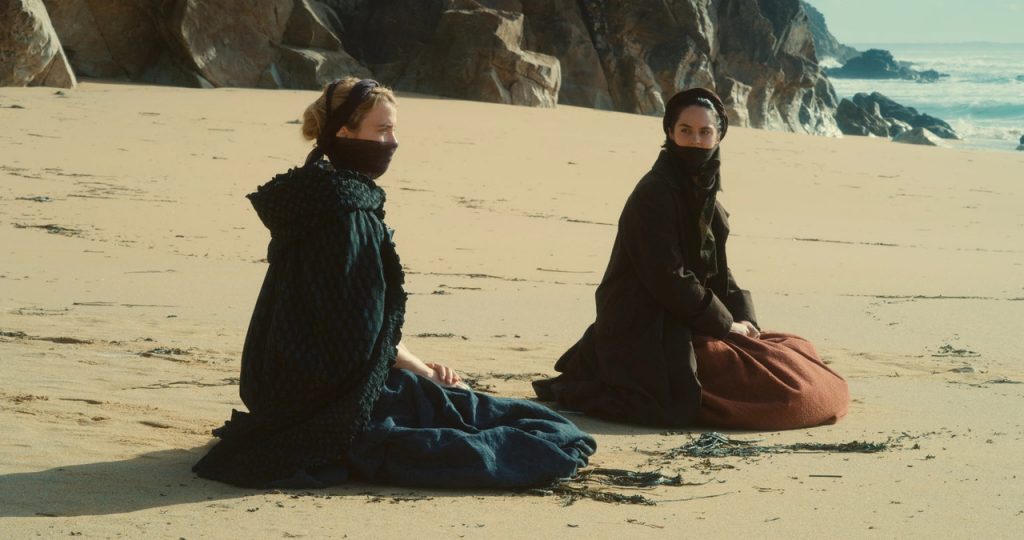 Imagem de Retrato de uma Jovem em Chamas. Nela, vemos Héloïse, à esquerda, com roupas azuis-escuras, e Marianne, à direita, com vestes vermelhas. Ambas usam um pano na boca para se cobrirem em um dia de ventania, e estão em uma praia, ajoelhadas na areia.