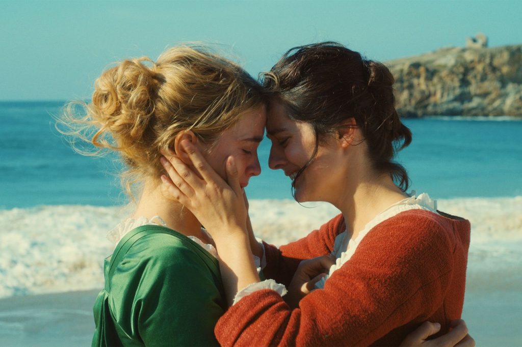 Imagem do filme "Retrato de uma Jovem em Chamas". Nela, vemos Héloïse e Marianne abraçadas em uma praia, em primeiro plano. Ambas estão chorando. Héloïse é loira, usa um vestido verde, e está à esquerda; enquanto Marianne, morena, segura o rosto de Héloïse com as duas mãos, e traja um vestido vermelho.