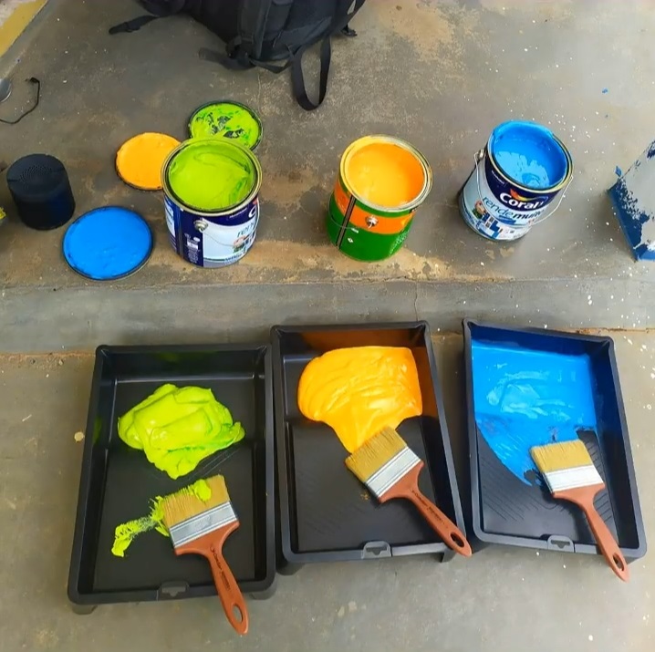 Foto de cima pra baixo das latas de tinta e pincel com as três cores escolhidas por Dequete: verde, amarelo e azul.