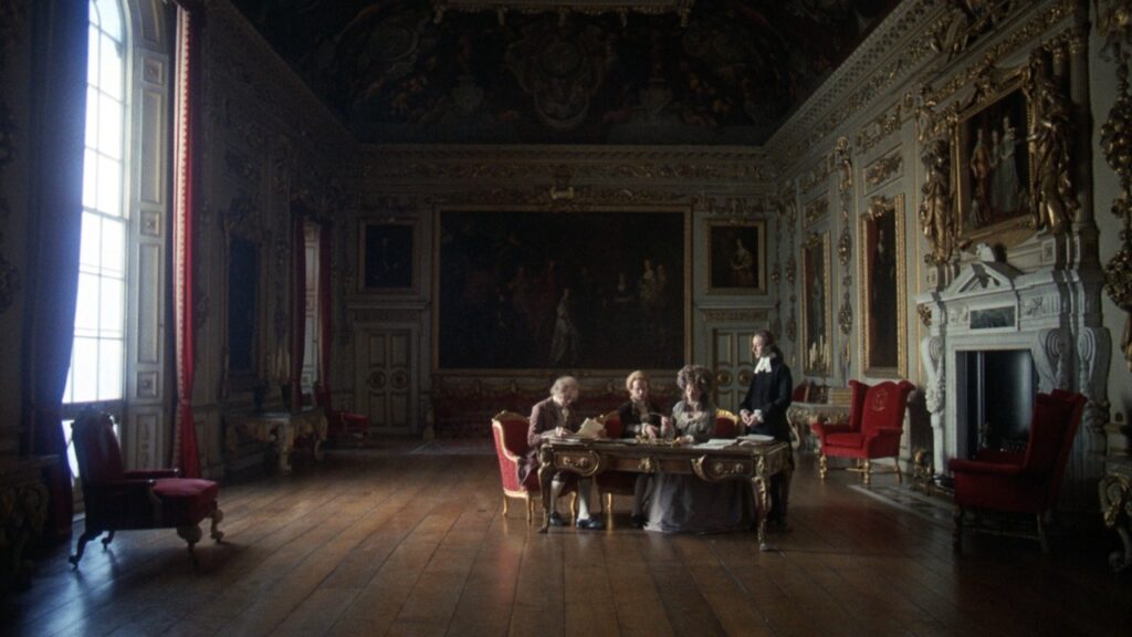 Imagem do filme "Barry Lyndon" (1975), de Stanley Kubrick. Nela, vemos três nobres sentados em uma mesa, enquanto um nobre está em pé, em um salão do século 18, adornado por quadros em todas as paredes. Os nobres estão longe na imagem, em um plano aberto.