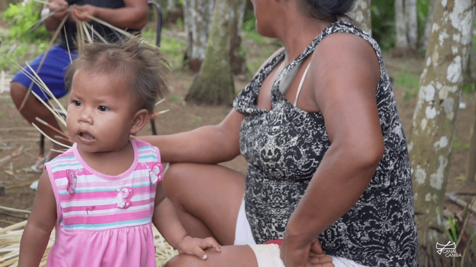 Criança indígena com trajes rosa. Ao seu lado, uma mulher com os braços apoiados nas coxas - Otageek