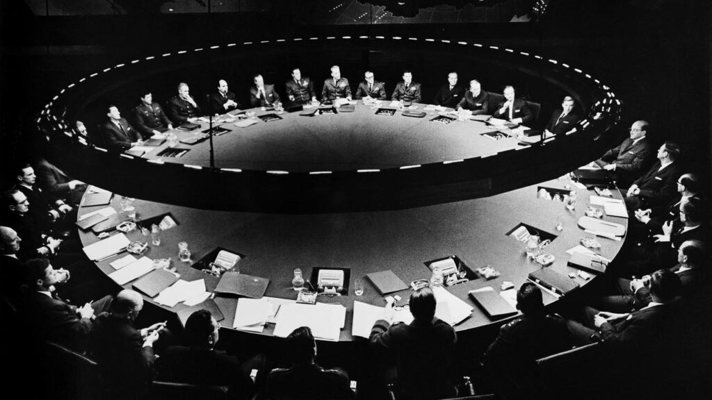 Imagem do filme "Dr. Fantástico" (1964), de Stanley Kubrick. Nela, vemos representantes dos EUA sentados em uma mesa redonda, com iluminação também redonda acima da mesa, em um ambiente chamado "sala de guerra".