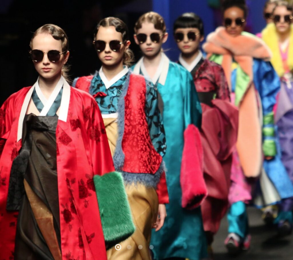 Imagem de um desfile de moda, onde várias modelos, todas de altura e cores de pele diferentes, mas com os mesmos óculos de sol redondos, desfilam com seus Hanboks - vestimenta tradicional coreana que se assemelha a um quimono