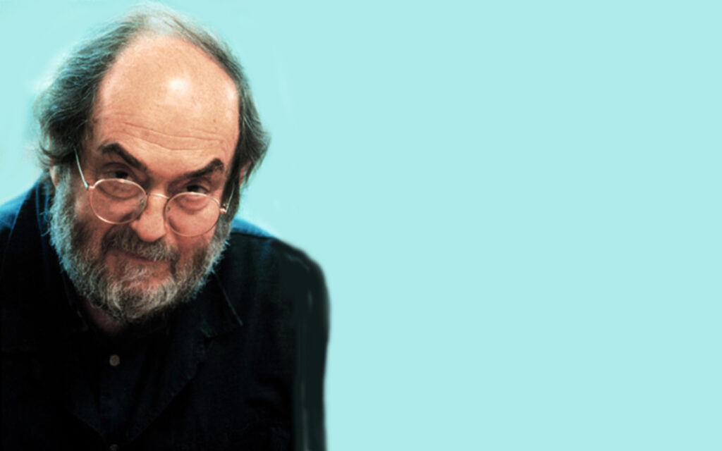 Foto do diretor estadunidense Stanley Kubrick. Nela, Kubrick, já de idade, meio careca e barbudo, usa óculos de grau redondos, um paletó preto e uma blusa de botões preta, e está alinhado à esquerda, de frente a um fundo azul-claro.