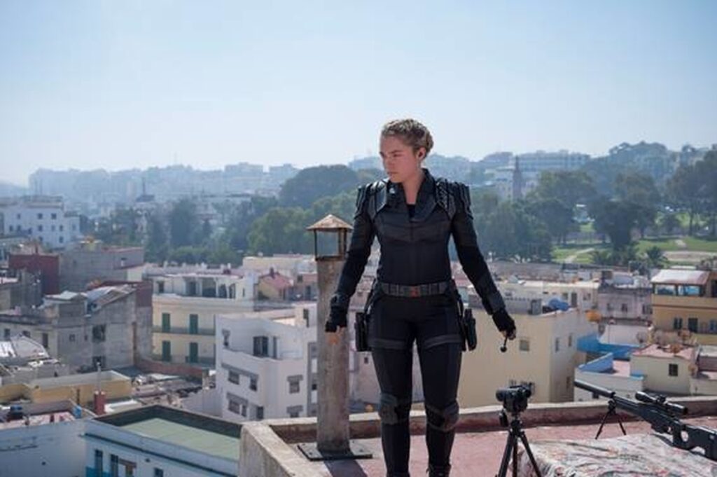 Imagem promocional do filme "Viúva Negra". Nela, vemos a personagem Yelena Belova, interpretada por Florence Pugh, em cima de um telhado na cidade de Budapeste, vestindo um traje preto e cercada por equipamentos militares.