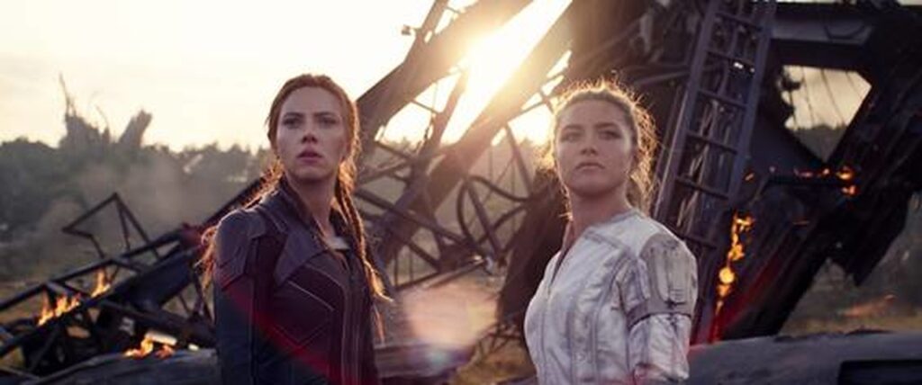 Imagem promocional do filme "Viúva Negra". Nela, mais uma vez, vemos as personagens Natasha e Yelena, respectivamente da esquerda para a direita e com um traje preto e um traje branco, na frente de destroços de uma explosão, olhando para o horizonte.