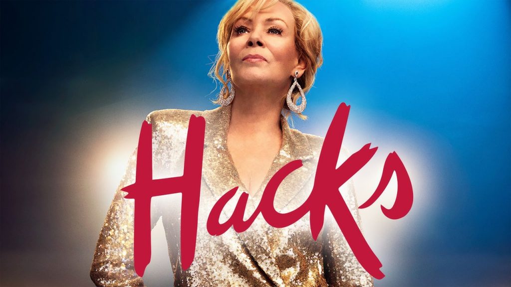 Na imagem temos a protagonista, Deborah Vance, em um terno dourado e a logo da série, "Hacks", em vermelho.