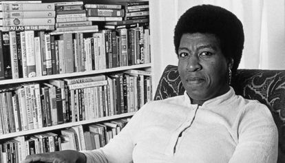 Escritora Octavia Butler, uma mulher negra, com cabelo curto, sentada em uma poltrona de frente a uma estnate de livros. 