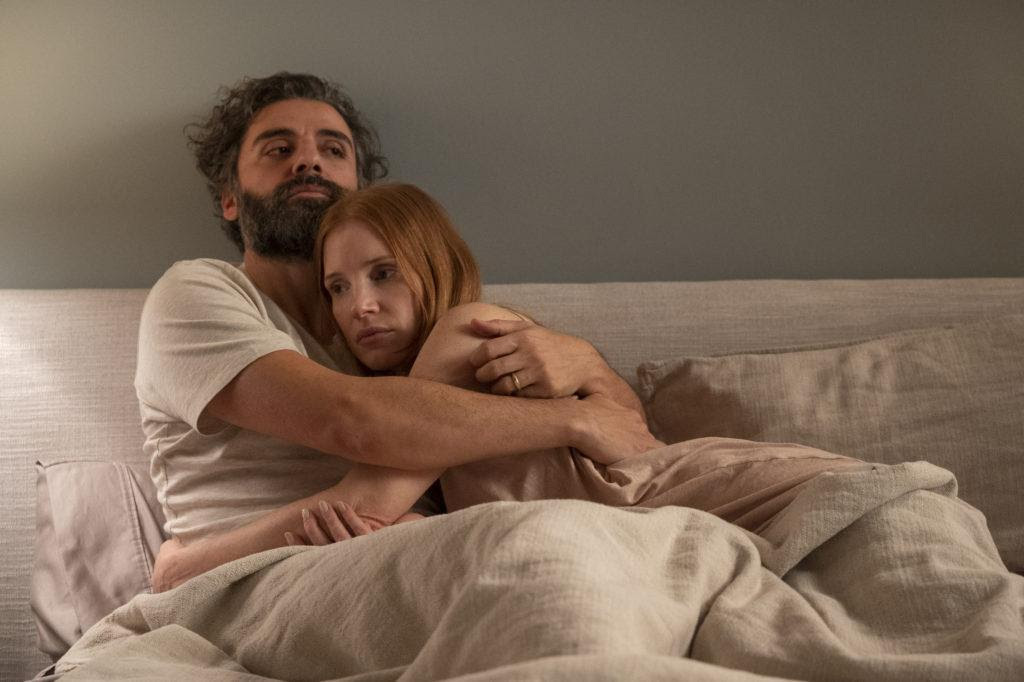 Homem latino de barba e cabelo escuros abraça mulher ruiva branca na cama.