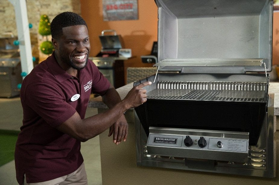 Imagem promocional de um filme. O ator Kevin Hart, um homem adulto, negro e com cabelo curto, está sorrindo enquanto aponta para uma máquina de cozinha que aparenta ser uma assadeira ou forno.