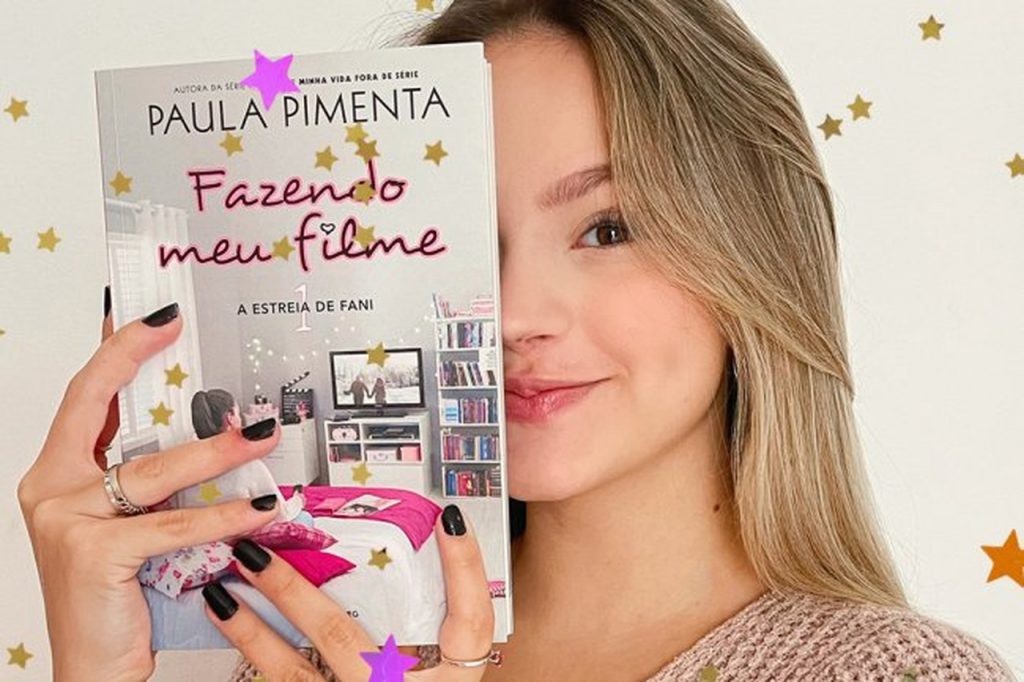 Foto da atriz Bela Fernandes, segurando o primeiro volume da série de livros "Fazendo Meu Filme". Bela interpretará Fani, a protagonista. Na foto, Bela segura o livro na altura do rosto, e o usa para tampar o lado direito (esquerda, para quem vê a foto) da face. Ao redor dela, em um fundo branco, há várias estrelinhas douradas.