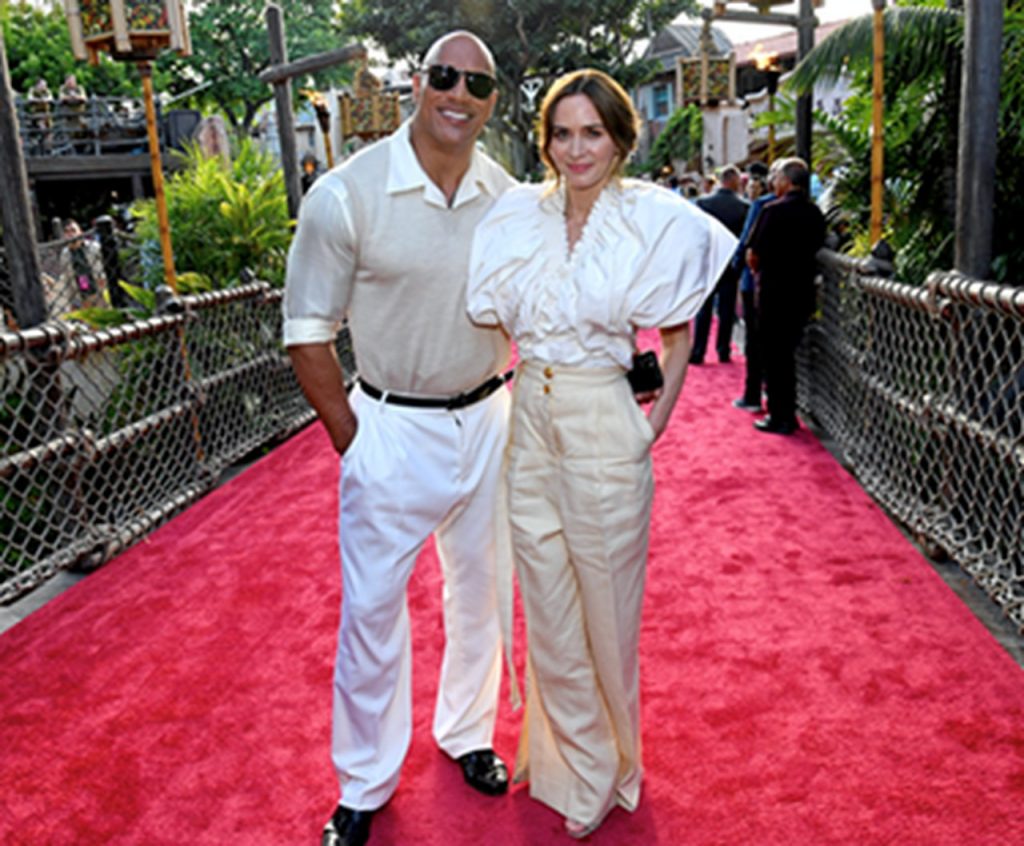 Imagem do tapete vermelho do filme "Jungle Cuise", na Disneyland Califórnia. Nela, vemos Dwayne Johnson e Emily Blunt, ambos com trajes brancos, em um tapete vermelho.
