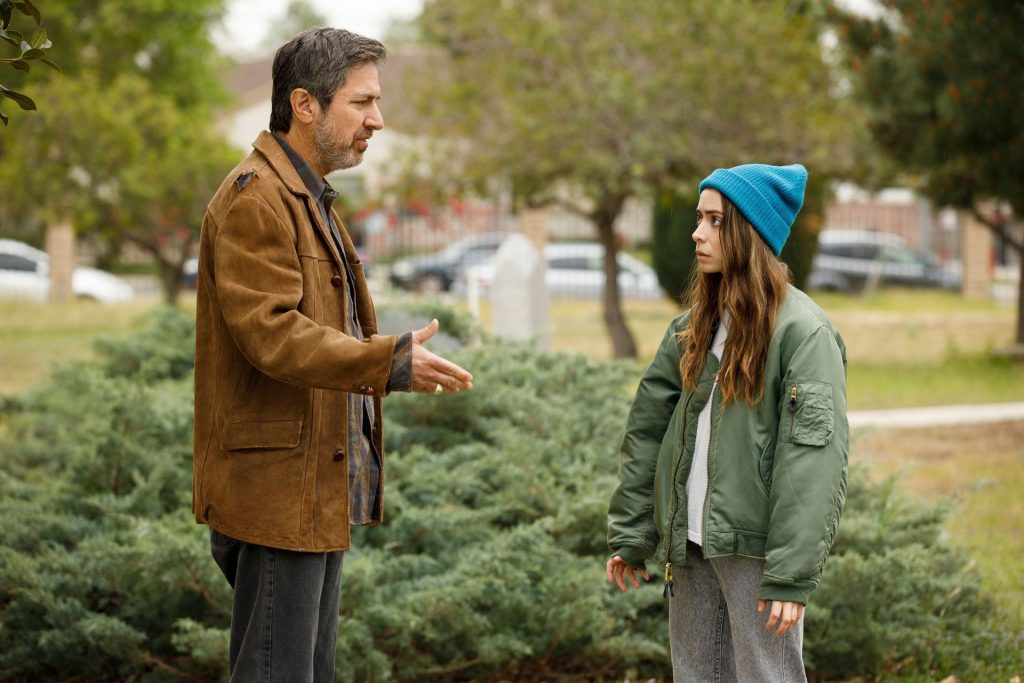 Um homem e uma mulher estão conversando. Ele está vestido com um casaco marrom e calça preta. Ela está de gorro azul, casaco verde e calça cinza. O cenário é um parque e um arbusto logo atrás deles. Na foto estão Hazel Green (Cristin Milliot) e Hebert Green (Ray Romano) e