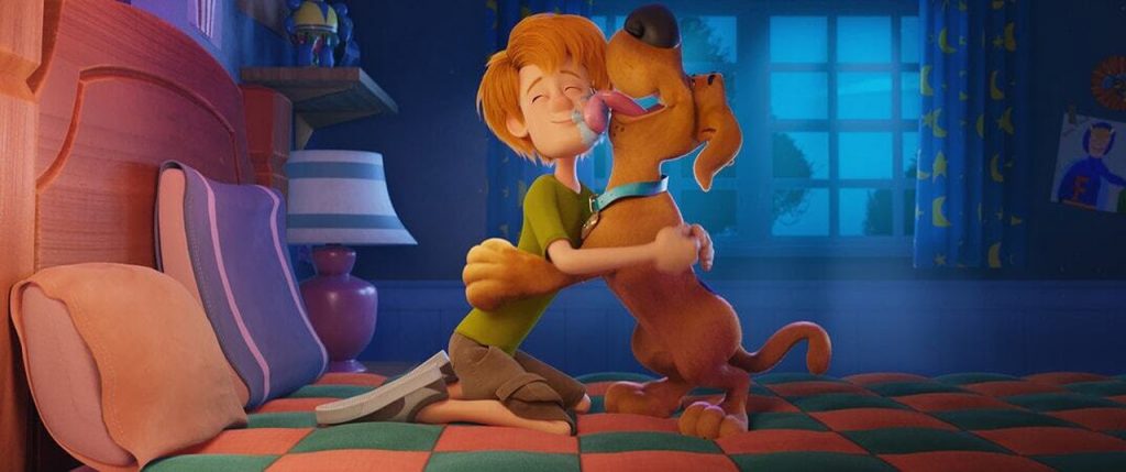 Imagem promocional do filme "Scooby! O Filme". Nela, vemos Salsicha e Scooby-Doo, dois companheiros, como pai e filho, abraçados em uma cama. Scooby está lambendo o rosto de Salsicha.