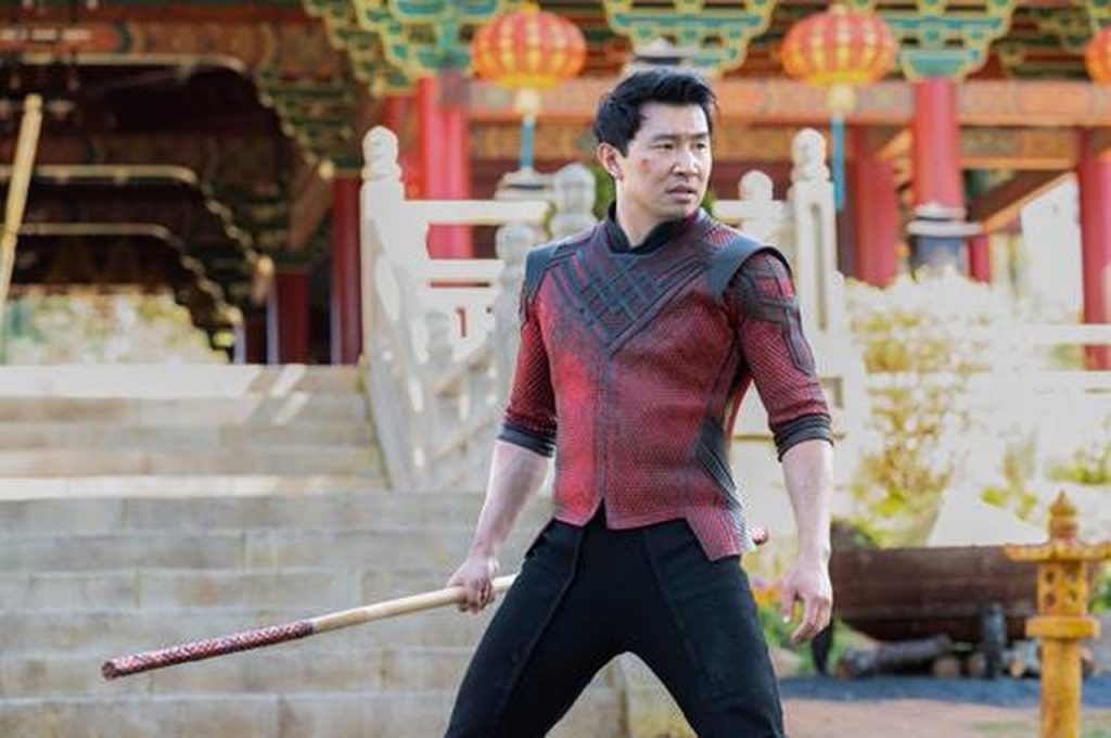 Imagem promocional do filme Shang-Chi e a Lenda dos Dez Anéis. Nela, o protagonista, Shang-Chi, interpretado por Simu Liu, está com uma camisa vermelha com adornos orientais, e segura um bastão. O cenário parece ser um templo, adornado com partes vermelhas, laranjas e verdes.