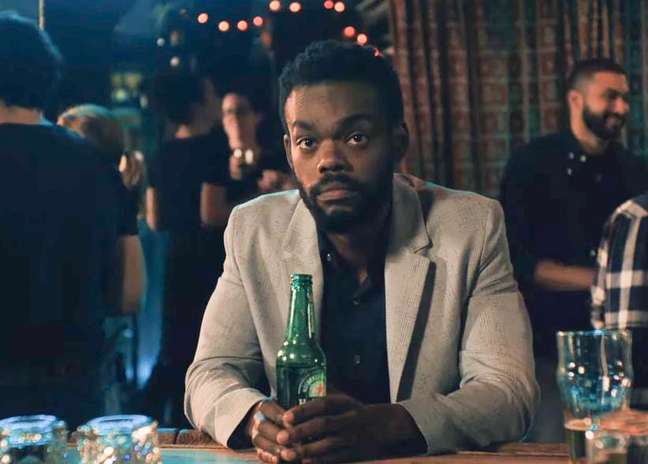homem negro de terno claro, sentado em uma mesa com uma garrafa de cerveja em mãos