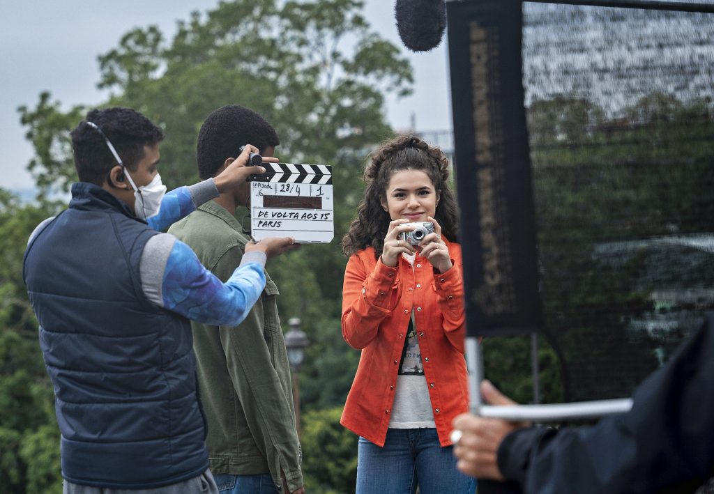 Foto dos bastidores da série De volta aos 15. A atriz Maísa está virada para a câmera, sorrindo de casaco laranja, calça jeans e segurando uma câmera fotográfica. Há dois membros da produção da série ao redor.