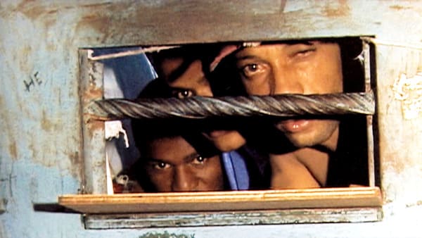 Três homens presos atrás de uma porta, olhando através da pequena janela com grades