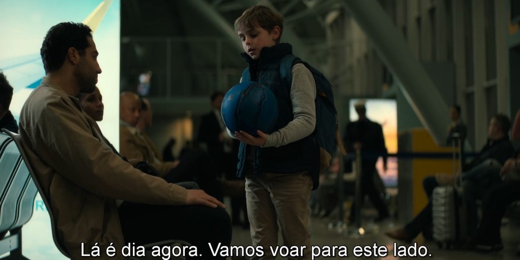 Elias explica para Farid sobre a rotação do planeta Terra no aeroporto, exemplificando em uma bola de basquete.