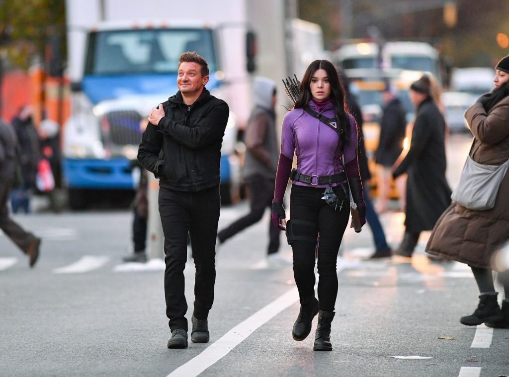 Imagem de bastidores da série "Gavião Arqueiro", da Disney+. Nela, vemos, em uma rua movimentada de Nova York, Clint Barton (Jeremy Renner), à esquerda, com jaqueta, calça e sapatos pretos, com uma mão no ombro e sorrindo, e à direita, vemos Kate Bishop (Hailee Steinfeld), com jaqueta roxa e calça preta, usando seus equipamentos de arco e flecha, com a cara fechada e segurando um livro.