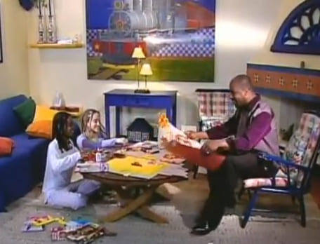 Duas meninas sentadas no chão de uma sala bem decorada recortando cartazes e uma homem negro sentado no sofá