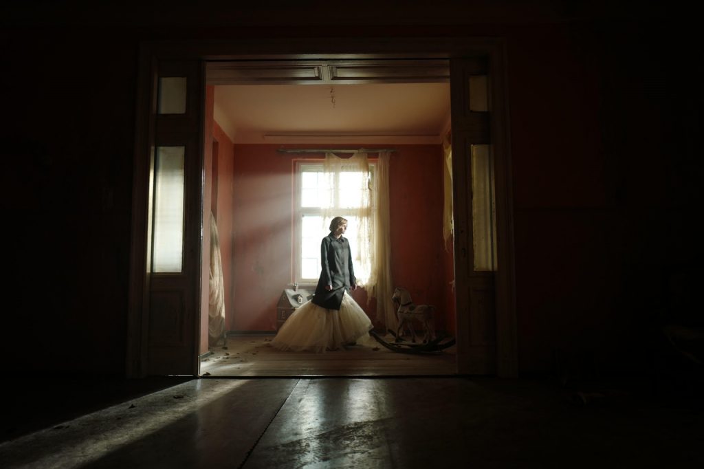 Imagem promocional do filme "Spencer", de Pablo Larraín. Nela, vemos a princesa Diana com um sobretudo e um vestido branco, dentro de um quarto abandonado, que parece ser o quarto de uma criança, a julgar pelos poucos objetos que ali estão, como um cavalinho de balanço.